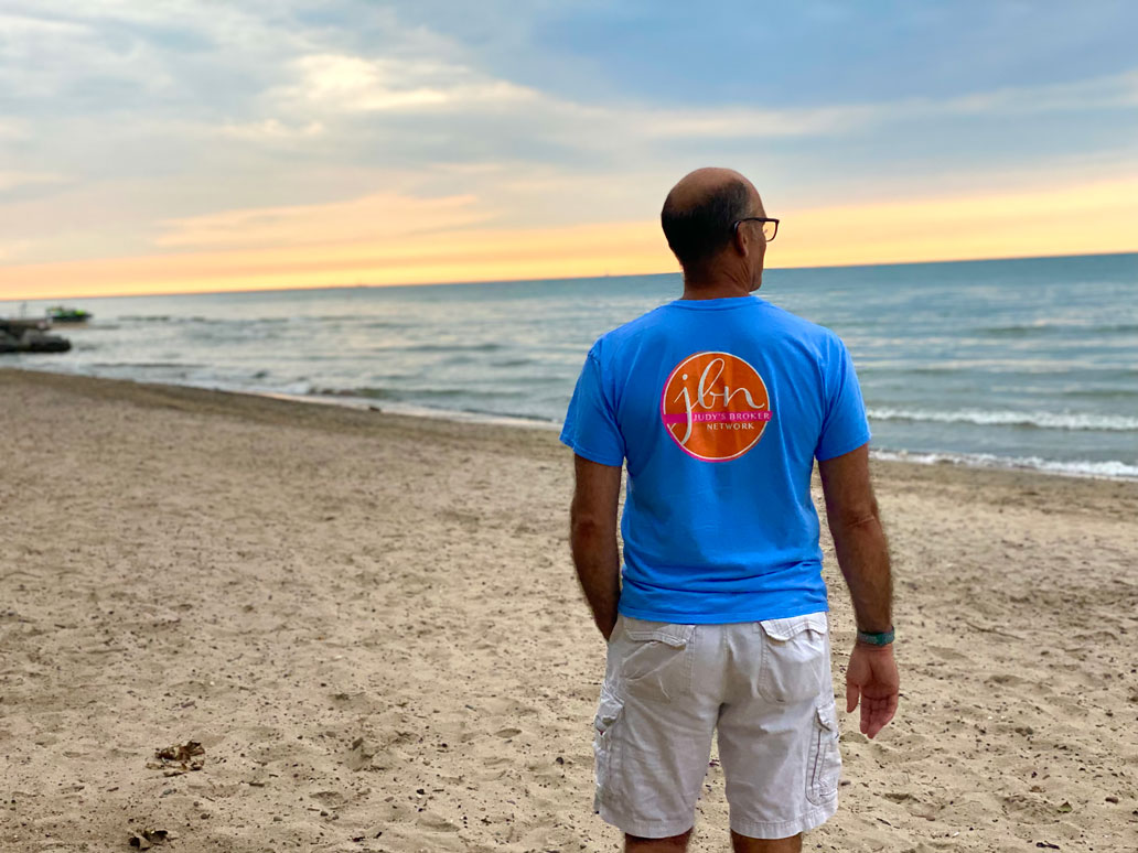 photo of man standing on beach at sunset wearing Judys Broker Network logo T-shirt